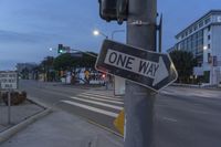 a one way sign on top of a metal pole, at a street intersection with cars