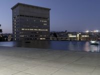 the walkway at the park leads to a lake and buildings in the background as evening falls