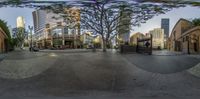 a panoramic photo taken in a city plaza with trees in it and several people on bikes and cars parked