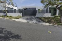 a street with trees, shrubs and bushes in front of a gray building with a gate