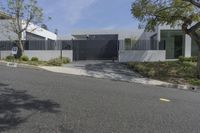 a street with trees, shrubs and bushes in front of a gray building with a gate