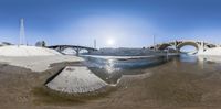 a fisheye view of a river and a bridge on the shore with a sand hill in the background