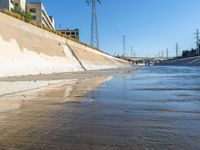 a river that is running on sand near a bridge with power lines and apartment buildings behind it