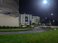 an empty street in front of some lit buildings at night with grass and a street light