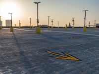 an empty parking lot during the sunset with broken yellow arrow marks on concrete and pole lights