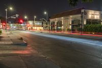 a street is shown with lights on and red stoplights overhead at night time, as the traffic goes in a blur