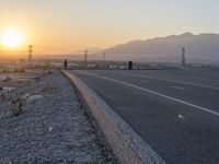 a motorcycle sits parked on a gravel path near the road with the sun setting behind it