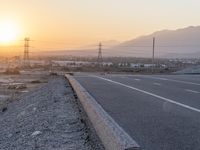 a motorcycle sits parked on a gravel path near the road with the sun setting behind it