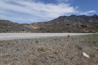 a dirt road with rocky terrain and mountains in the distance, with bushes on either side of the road