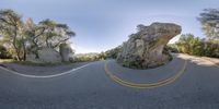 an asphalt road that has a rock in the middle with trees in the background, in front of some rocks and a sky