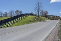 a motorcycle is riding down a rural road in front of a fence and green hill