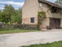 a brown brick building with wooden doors in the grass near a street corner with a bench and picnic table