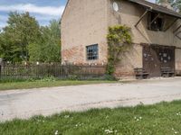 a brown brick building with wooden doors in the grass near a street corner with a bench and picnic table