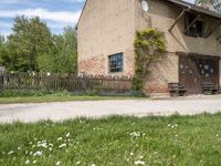 a brown brick building with wooden doors in the grass near a street corner with a bench and picnic table