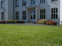 a lawn area in front of a white, four - story building with red flowers