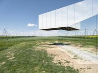 a mirror shaped sculpture is surrounded by grass and power lines and wind turbines on a clear day