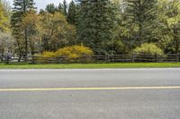 some trees in the background and a fence around one side of the road and a sign in front of it with yellow marking on it