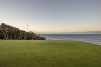 there is a lush green lawn overlooking the ocean at sunset and a few trees in the distance