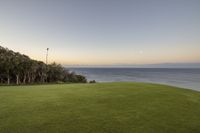 there is a lush green lawn overlooking the ocean at sunset and a few trees in the distance