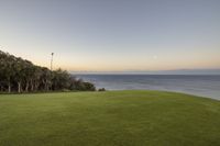 there is a lush green lawn overlooking the ocean at sunset and a few trees in the distance