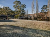 a park bench sits beside water and trees as a person stands at a picnic table in a frosty park area