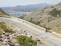 there is a person standing on the road by the water and mountains behind them with a vehicle on the road