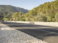 a paved highway is surrounded by rocks and forest on a sunny day from the side