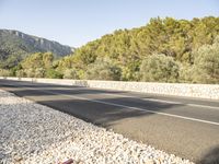 a paved highway is surrounded by rocks and forest on a sunny day from the side