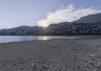 a horse is sitting in the sand by water and mountains in a blue sky and white clouds