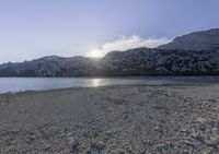 a horse is sitting in the sand by water and mountains in a blue sky and white clouds