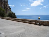 a paved road is next to the shore with a stone fence and a large cliff