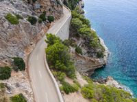 two people on bikes riding down a narrow road near the ocean, in front of cliffs