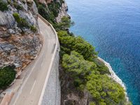 two people on bikes riding down a narrow road near the ocean, in front of cliffs