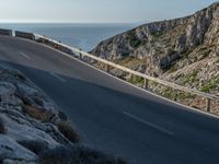 a person riding a bike down a road by the water and cliff side, looking back