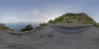 a curved road with rock, trees and sky in the background and mountain backdrop on either side of road