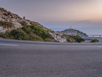 a road at the edge of a cliff next to the ocean at dusk, on which there is a light tower