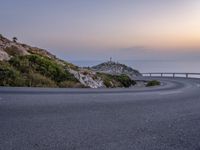 a road at the edge of a cliff next to the ocean at dusk, on which there is a light tower