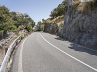 a motorcycle is traveling down the mountain pass of a steep cliff lined road with bushes, grass and trees