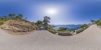 a street in front of a cliff next to water and mountains with the sun in the sky