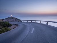 a curved road is shown next to the ocean at sunset on a mountaintop with a lighthouse in the distance