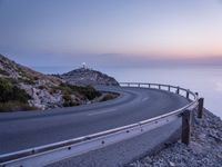 a curved road is shown next to the ocean at sunset on a mountaintop with a lighthouse in the distance