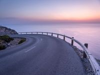 a curved road is shown next to the ocean at sunset on a mountaintop with a lighthouse in the distance