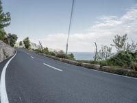 a winding road on a clear day with the ocean visible in the background, with bushes and trees around the curve,