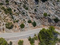 an aerial shot of a motorcycle driving by rocks and cliffs, near a pine tree lined road