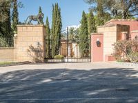 a driveway with some trees and some stone walls and a fence and gates with a statue of a horse