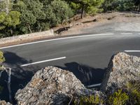 a motorcycle is driving along the empty road near the trees and rocks with the shadow on the pavement