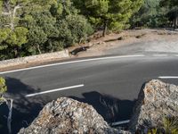 a motorcycle is driving along the empty road near the trees and rocks with the shadow on the pavement