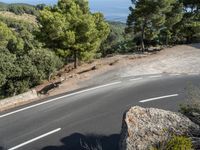 a motorcycle is driving along the empty road near the trees and rocks with the shadow on the pavement