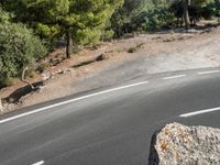 a motorcycle is driving along the empty road near the trees and rocks with the shadow on the pavement