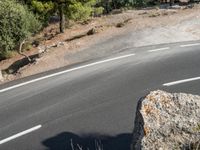 a motorcycle is driving along the empty road near the trees and rocks with the shadow on the pavement